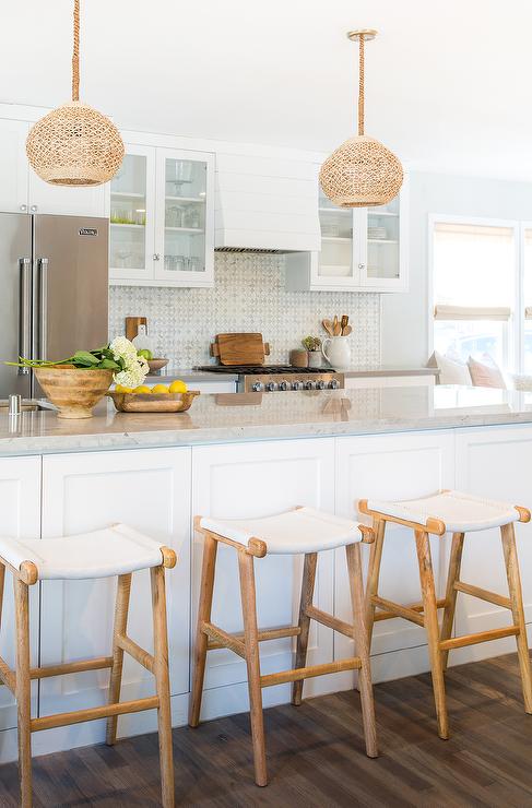 White and gray kitchen features two woven dome pendant lights hanging over a long white center island lined with backless wood and white leather bar stools White and gray kitchen features two woven dome pendant lights hanging over a long white center island lined with backless wood and white leather bar stools
